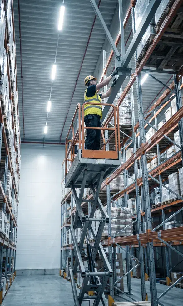 A specialist in a high-visibility vest and helmet on a scissor lift platform, inspecting an upper beam connection of warehouse racking at height